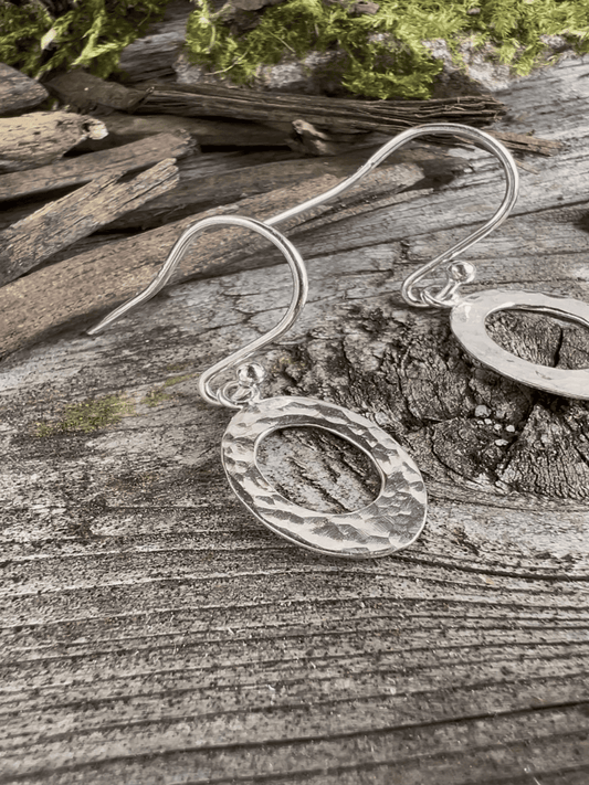 Close-up of handmade sterling silver textured oval earrings on weathered wood, highlighting the hammered surface texture on the oval frame
