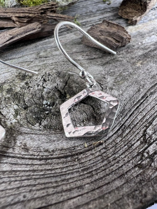 Close-up of handmade sterling silver textured pentagon earrings on weathered wood, highlighting the hammered geometric frame