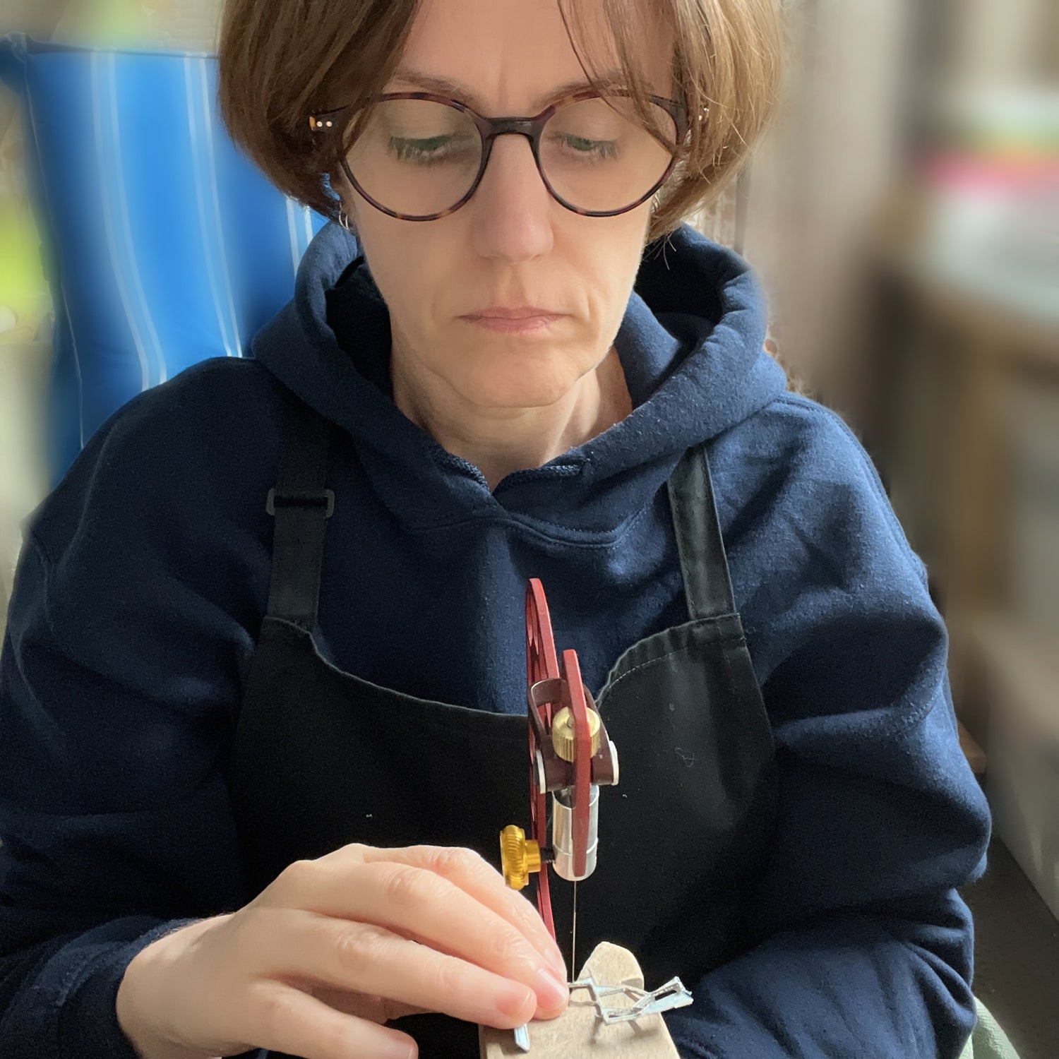 Person wearing glasses and a blue apron piercing silver at the bench peg.