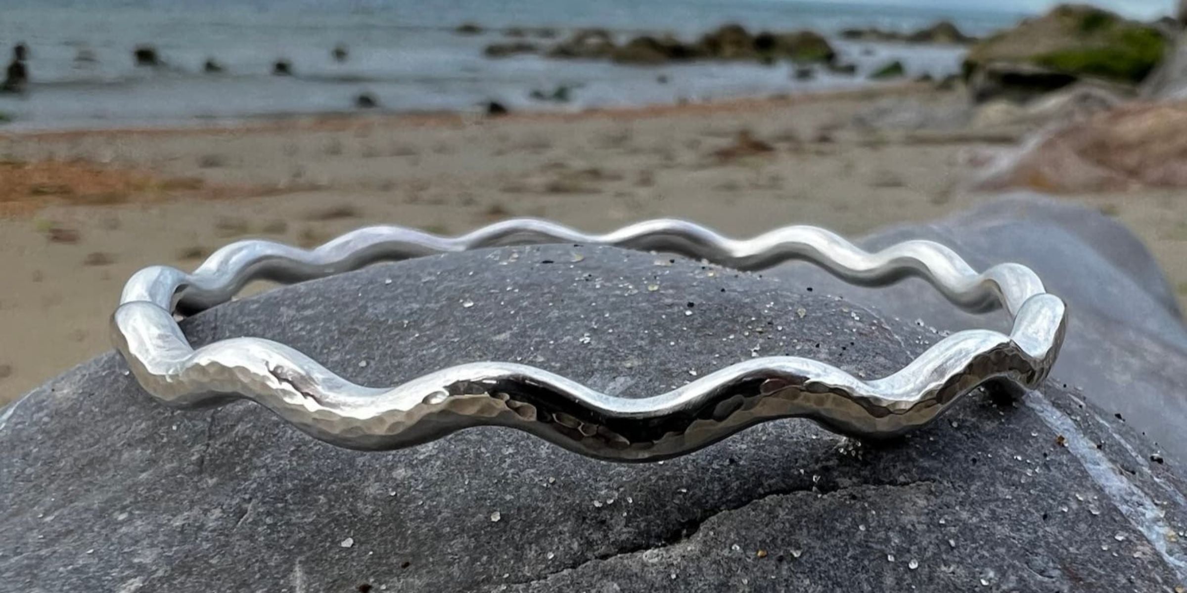 Sterling Silver bracelet with wavy design on a rock by the sea, with text about handcrafted silver jewellery.