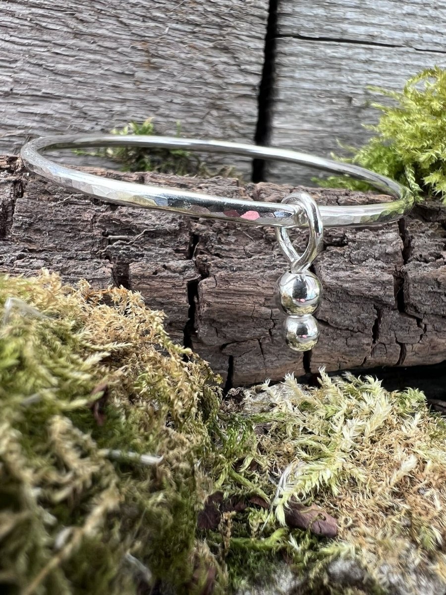 Close-up of a handmade sterling silver bauble charm bangle on mossy wood, highlighting the small bead charms