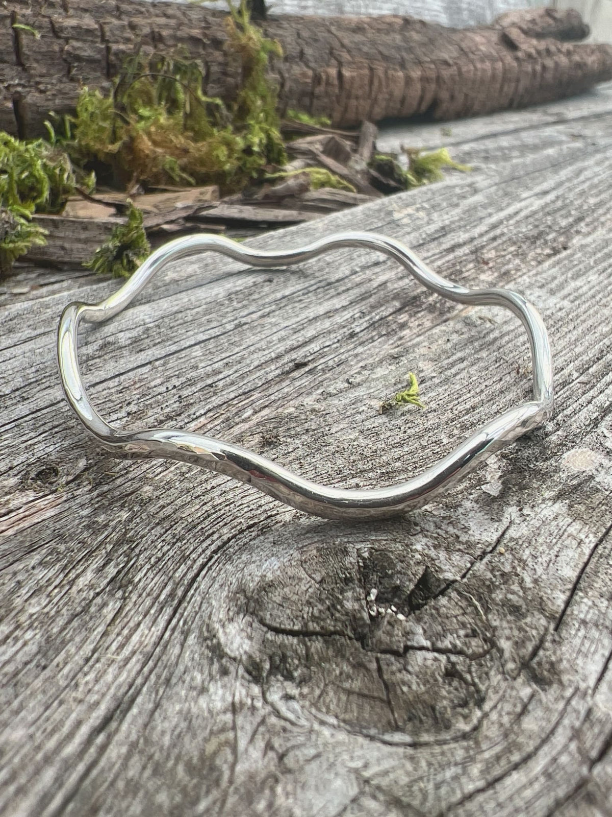 Handmade sterling silver gentle wave bangle 2.5mm on mossy driftwood, overhead angle showing the full wave outline