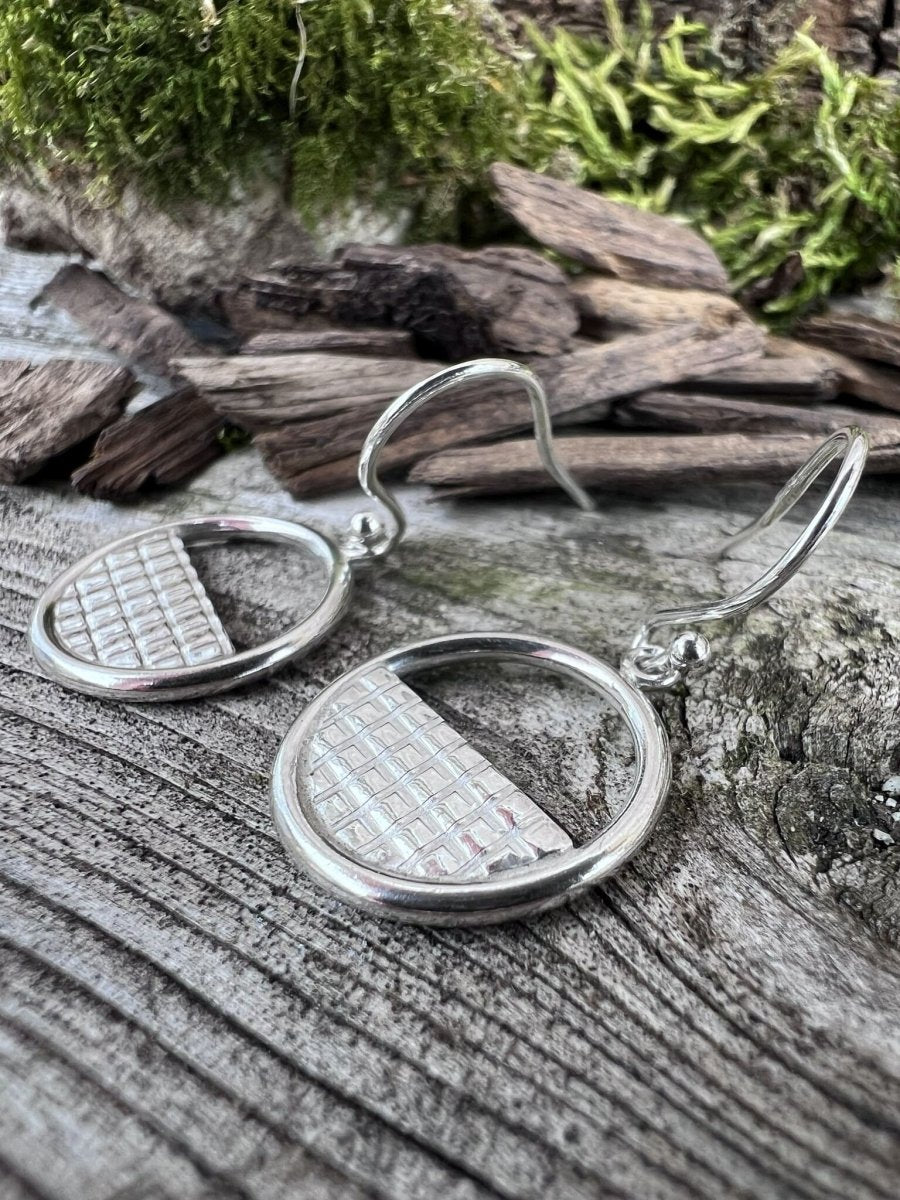 Close-up of handmade sterling silver lattice halo earrings on mossy weathered wood, highlighting the fine grid lattice detail inside the circle frame