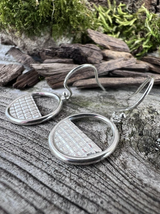 Close-up of handmade sterling silver lattice halo earrings on mossy weathered wood, highlighting the fine grid lattice detail inside the circle frame