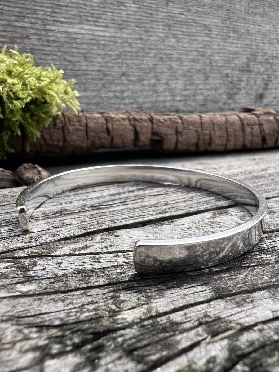 Recycled sterling silver Solstice sleek torque bangle lying flat on weathered wood with green moss