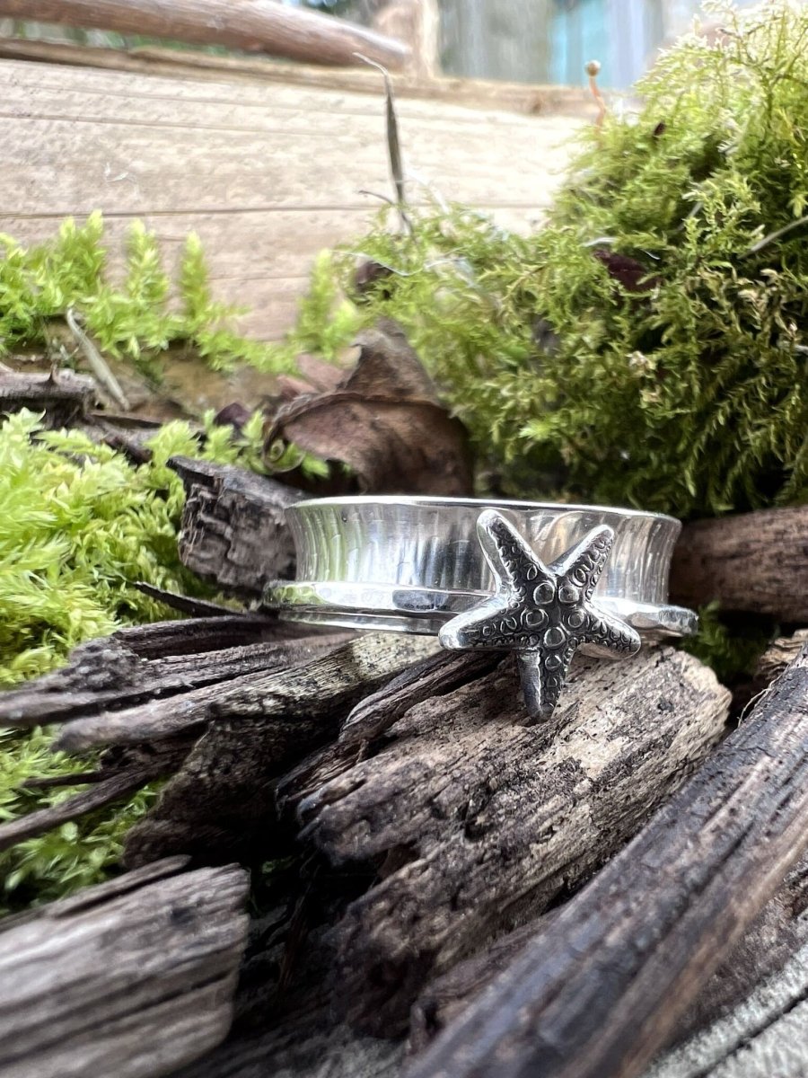 Handmade recycled silver spinner ring with starfish on wood chips surrounded by moss, viewed from a wider angle