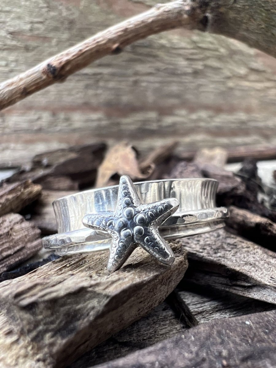 Close-up of a handmade sterling silver spinner ring with a starfish charm on wood chips, highlighting the 3D starfish and spinning band