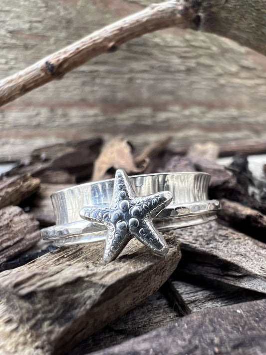 Close-up of a handmade sterling silver spinner ring with a starfish charm on wood chips, highlighting the 3D starfish and spinning band
