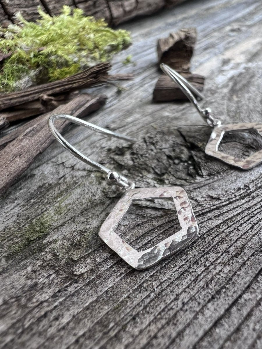 Handmade sterling silver textured pentagon earrings on weathered wood with foliage, photographed from an angle showing the open frame
