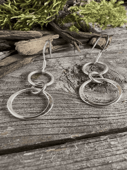 Handmade sterling silver unity drop earrings on weathered wood with foliage, photographed from an angle showing pendant depth