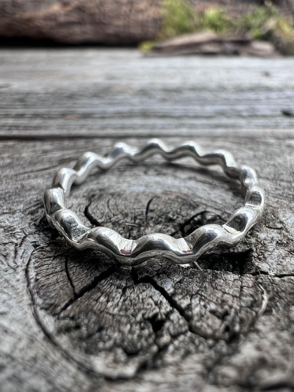 Close-up of a handmade sterling silver wave ring on driftwood, highlighting the rippled wave edge along the band